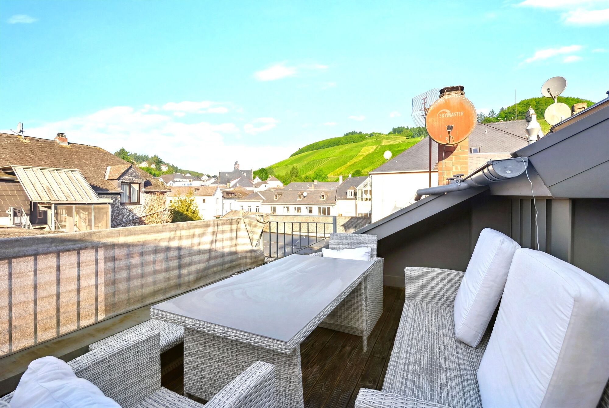 Graue Lounge-Möbel auf einer Dachterrasse, im Hintergrund grüne Weinberge und Hausdächer. Graue Lounge-Möbel auf einer Dachterrasse, im Hintergrund grüne Weinberge und Hausdächer.