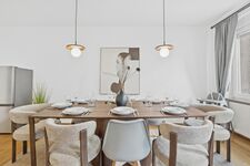 Langer Holztisch mit grau-beigen Polsterstühlen, zwei Pendelleuchten, abstraktes Kunstbild, Hochstuh Langer Holztisch mit grau-beigen Polsterstühlen, zwei Pendelleuchten, abstraktes Kunstbild, Hochstuh