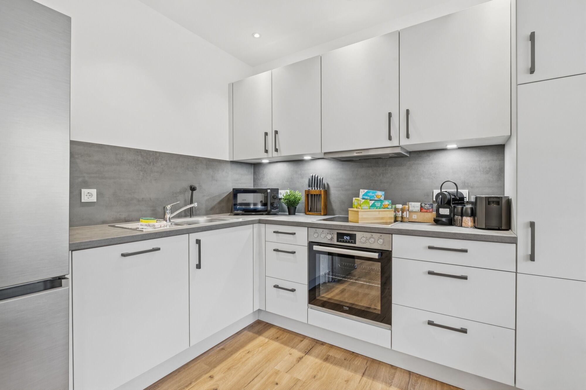 Weiße L-Küche mit grauer Arbeitsplatte, Backofen, Mikrowelle, Fenster mit Grünblick. Weiße L-Küche mit grauer Arbeitsplatte, Backofen, Mikrowelle, Fenster mit Grünblick.