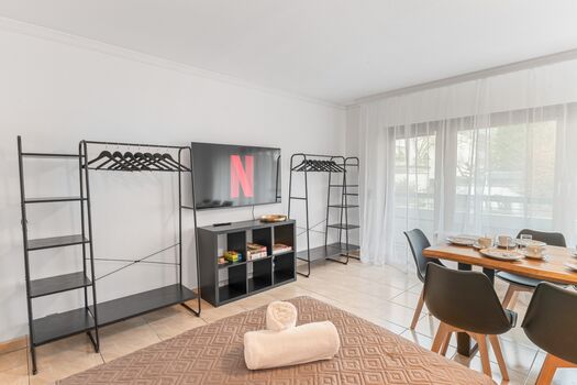 Helles Wohn- und Esszimmer mit 55 Zoll Smart-TV. Offene Kleiderständer und Regale bieten ausreichend Stauraum für längere Aufenthalte. Viel Tageslicht durch große Fensterfront.